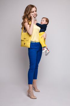 Side view of pretty mother walking with little kid and taking selfie. Young woman keeping child on hands and posing on isolated background in studio. Concept of childhood and family outfit.