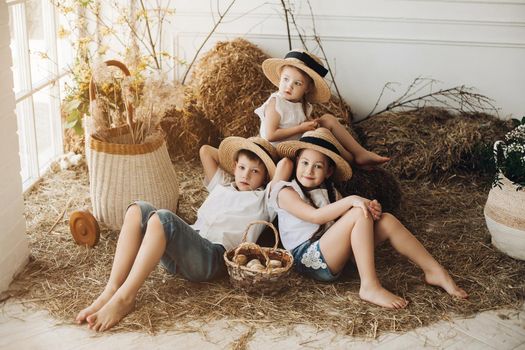 Cute sisters and brother wearing hay hats, white shirts and shorts resting and lying on hay in village. Little children looking at camera, smiling and posing. Concept of family and childhood.