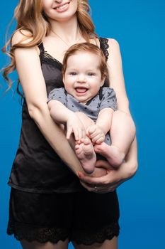 Front view of positive little baby laughing with mother in studio. Beautiful woman in black pajamas keeping cute daughter on hands. looking at camera and posing on blue isolated background.