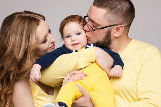 Front view of cheerful family in yellow outfit posing on grey isolated background. Pretty young mother, cheerful father and little baby looking at camera and smiling. Concept of love.