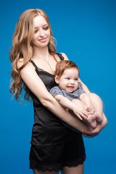 Front view of positive little baby laughing with mother in studio. Beautiful woman in black pajamas keeping cute daughter on hands. looking at camera and posing on blue isolated background.