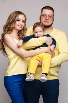 Front view of cheerful family in yellow outfit posing on grey isolated background. Pretty young mother, cheerful father and little baby looking at camera and smiling. Concept of love.