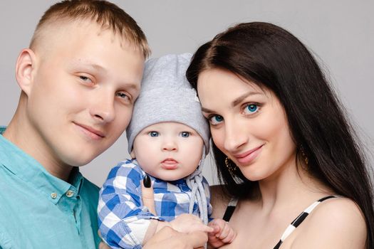 Studio headshot of cute baby boy in casual hat and checked shirt with his beautiful mother and handsome father. Family smiling at camera together.