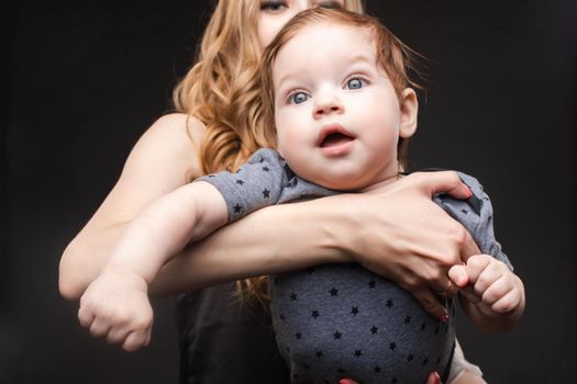 Front view of positive little baby laughing with mother in studio. Beautiful woman in black pajamas keeping cute daughter on hands. looking at camera and posing on black isolated background.