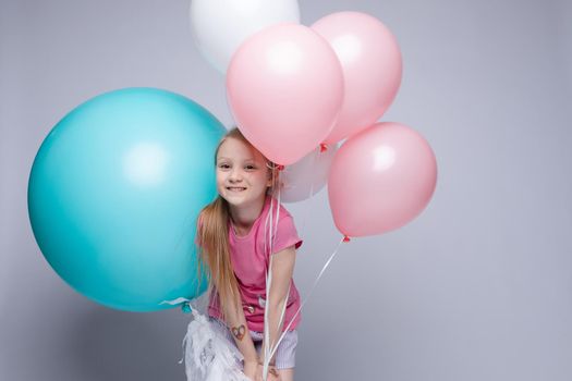 Studio portrait of lovely little girl with red or fair hair and freckles in pink t-shirt and shorts posing with toothy smile with pink, blue and white air balloons against grey background.