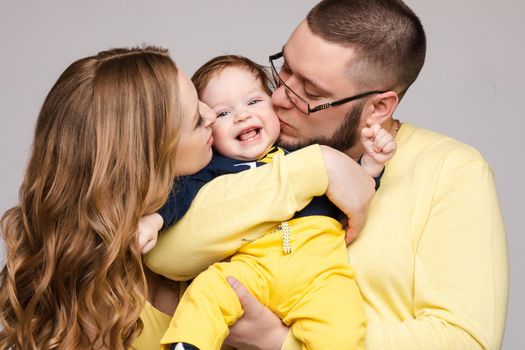 Portrait of happy family from three people wearing same coloured clothes. Blonde beautiful mother smiling and holding on hands surprised child. Handsome father in glasses hugging lovely wife and son.