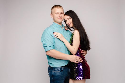 Beautiful young pair embracing each other in studio. Attractive woman in bright dress and man in blue shirt and jeans looking at camera, smiling and posing on isolated background. Concept of love.