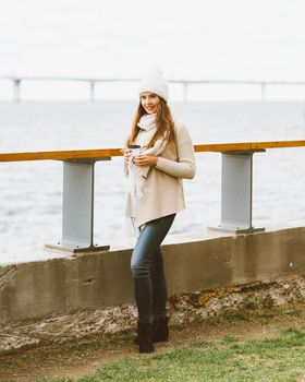 Beautiful young girl drinking coffee, tea from plastic mug in autumn, winter. A woman with long hair stands on waterfront on Baltic sea in port and waiting for ferry, heated by a hot drink, copy space