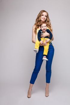 Young and fit woman in casual clothes holding little kid. Beautiful mother in yellow blouse and blue pants posing from back with her lovely son. Blonde mom on heels looking and happy smiling child.