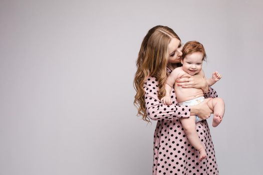 Front view of young mother keeping little child on hands and laughing on isolated background. Cheerful family posing and looking at camera in studio. Concept of happiness and childhood.