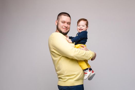 Portrait of happy father in yellow sweater posing with his lovely smiling child. Athletic and young men with beard holding red haired little boy. Handsome brunette dad hugging sweet and cute son.