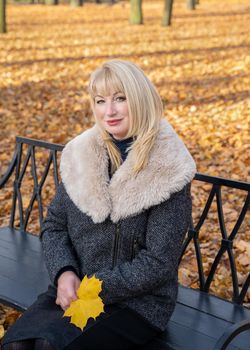 Happy blonde mature woman is sitting on bench in autumn park and thinking. Beautiful woman is relaxing in nature on a sunny day. Portrait of middle aged woman smiling and daydreaming.
