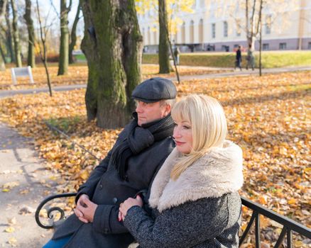 Closeup portrait of happy blonde mature woman and beautiful middle-aged brunette, looking away. Loving couple of 45-50 years old sitting on bench in autumn park in warm clothes, enjoys life