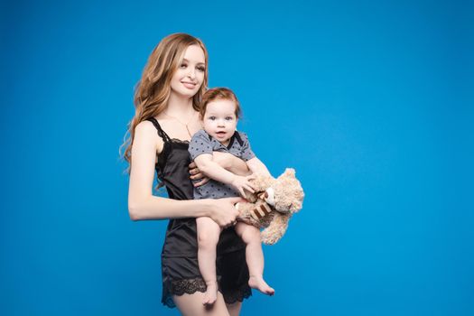 Front view of positive little baby laughing with mother in studio. Beautiful woman in black pajamas keeping cute daughter on hands. looking at camera and posing on blue isolated background.