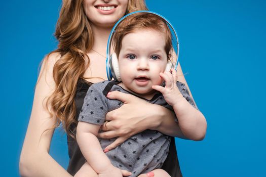 Studio portrait of cute little boy listening to music in headphones sitting on his mother s arms. Isolate on blue.