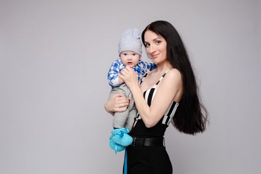 Happy beautiful mother holding child on hands and smiling. Side view of brunette woman with long hair together with baby in blue shirt and grey pants. Young parent hugging little son in funny socks.