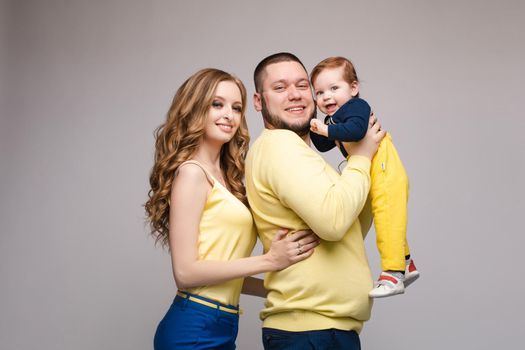 Front view of cheerful family in yellow outfit posing on grey isolated background. Pretty young mother, cheerful father and little baby looking at camera and smiling. Concept of love.