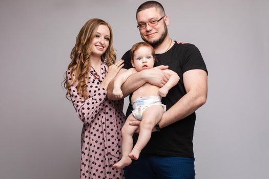 Studio portrait of beautiful young family of three. Mother, father and a baby boy smiling happily at camera. They are isolated on grey background.