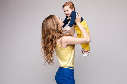 Young and fit woman in casual clothes holding little kid. Beautiful mother in yellow blouse and blue pants posing from back with her lovely son. Blonde mom on heels looking and happy smiling child.