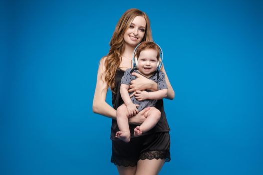 Front view of positive little baby laughing with mother in studio. Beautiful woman in black pajamas keeping cute daughter on hands. looking at camera and posing on blue isolated background.