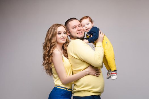 Portrait of happy family from three people wearing same coloured clothes. Blonde beautiful mother smiling and holding on hands surprised child. Handsome father in glasses hugging lovely wife and son.