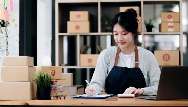 A portrait of a young Asian woman, e-commerce employee sitting in the office full of packages in the background write note of orders and a calculator, for SME business, e-commerce and delivery business..