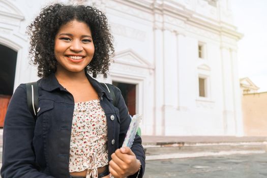 Curly-haired mestizo university student with a backpack and a notebook in her hand smiling and looking at the camera outdoors in the central park of leon nicaragua..