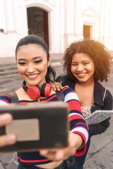 Vertical photo of two multiracial Latina students taking a self-portrait with their cell phone smiling outdoors in a public square in Leon
