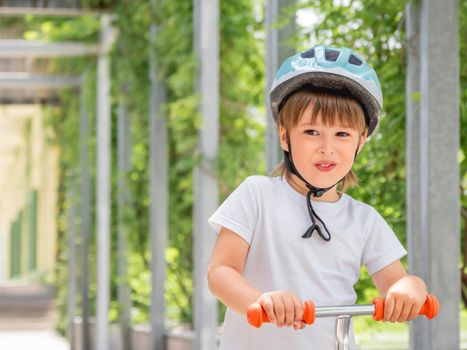 Little boy rides kick scooter in skate park. Portrait of kid in helmet on green leaves background. Training to skate at summer.