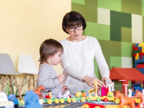 Toddler plays with colorful toy blocks while his mother or babysitter. Little boy stares on toy constructor. Interior of kindergarten or nursery.