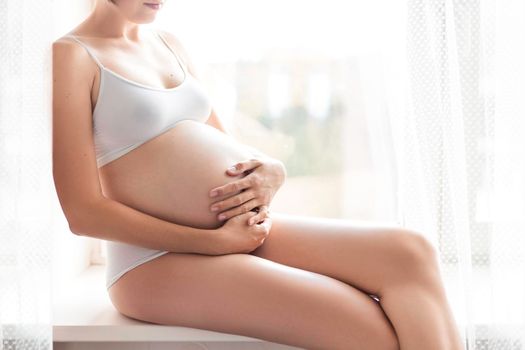 Pregnant woman in white underwear sitting on window sill. Young woman expecting baby. Cozy happy background in sunny morning.