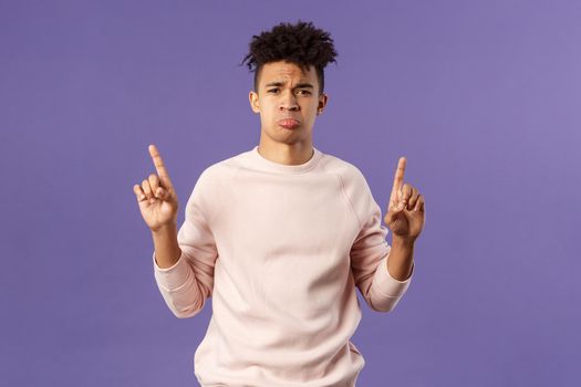 Portrait of gloomy, pouting frowning hipster guy dont have something he wants, pointing fingers up at super cool expensive thing, asking for it, trying receive compassion, purple background.