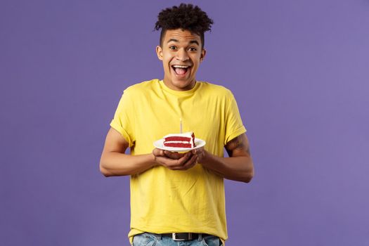 Happy birthday to me. Portrait of upbeat, excited hispanic man with dreads celebrating b-day, holding plate cake with lit candle, smiling amused, making wish, purple background.