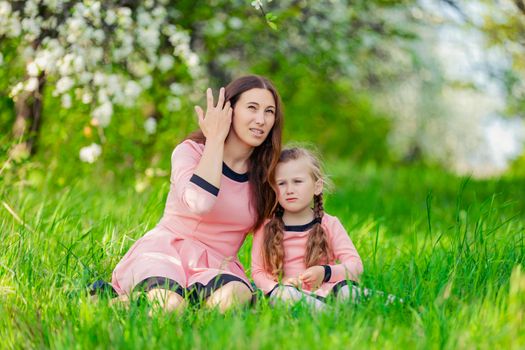mother and daughter sit in the green grass against the backdrop of blooming apple trees