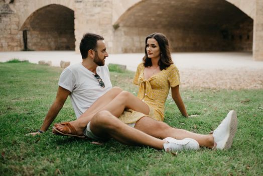A brunette girl in a yellow dress is sitting on the legs of her boyfriend on the grass in old Spain town. A couple of tourists on a date in the Valencian park.