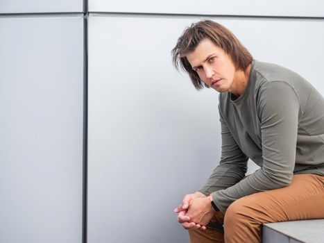 Millennial man with long hair and tense look sits near grey wall. Serious man in earth toned casual clothes is staring in camera. Copy space.