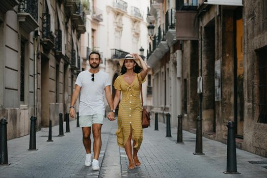 A girl in a hat and a yellow dress with a plunging neckline and her boyfriend with a beard are walking holding each other's hand in old Spain town. A couple of tourists on a date in Valencia.