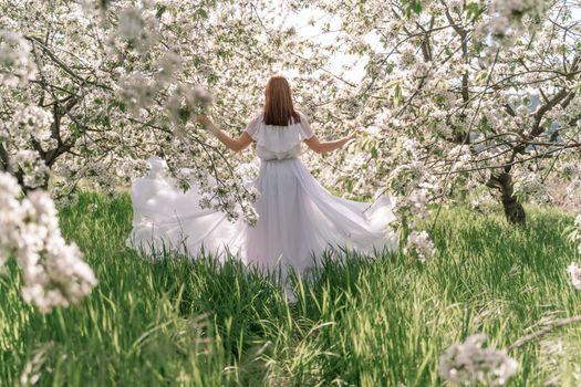 Fantasy woman in long white elegant fashion long dress walks in green spring blossom cherry garden. Happy cheerful girl princess bride. Skirt fabric flies flowing waving in wind motion