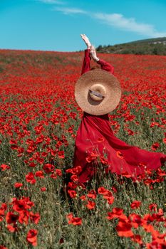 Young woman stands with her back in a long red dress and hat, posing on a large field of red poppies.