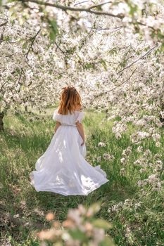 Fantasy woman in long white elegant fashion long dress walks in green spring blossom cherry garden. Happy cheerful girl princess bride. Skirt fabric flies flowing waving in wind motion