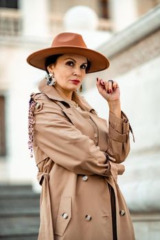 Outdoor fashion portrait of young elegant fashionable brunette woman, model in stylish hat, choker and light raincoat posing at sunset in European city