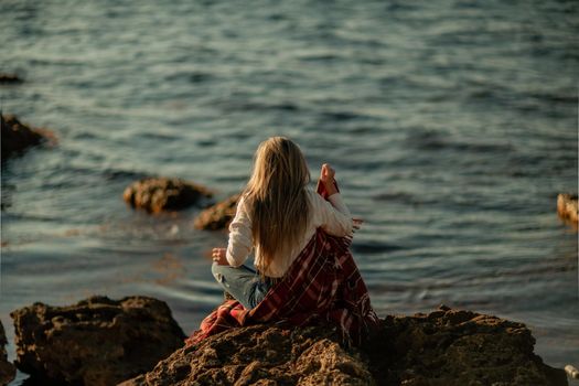 Attractive blonde Caucasian woman enjoying time on the beach at sunset, sitting in a blanket and looking to the side, with the sunset sky and sea in the background. Beach vacation