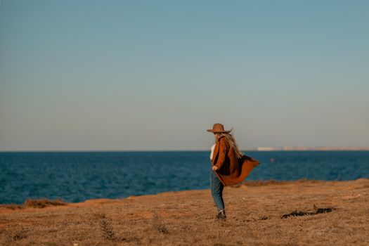 A woman walking along the coast near the sea. An elegant lady in a brown coat and a hat with fashionable makeup walks on the seashore.