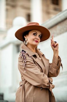 Outdoor fashion portrait of young elegant fashionable brunette woman, model in stylish hat, choker and light raincoat posing at sunset in European city
