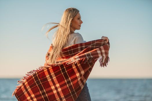 Attractive blonde Caucasian woman enjoying time on the beach at sunset, walking in a blanket and looking to the side, with the sunset sky and sea in the background. Beach vacation.
