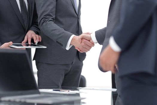 Two cheerful businessmen shaking hands with smile at office with their coworkers.