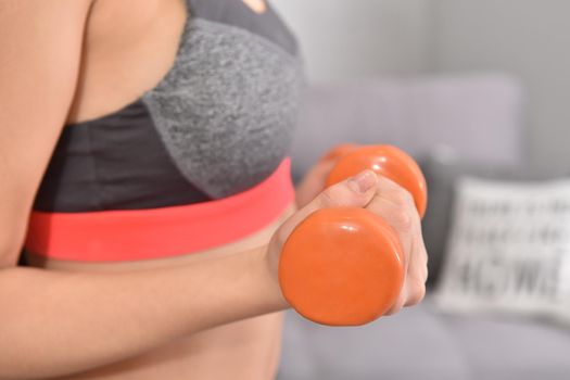 Young asian brunette woman in sportwear making home workout with two orange dumbbells. Soft focus