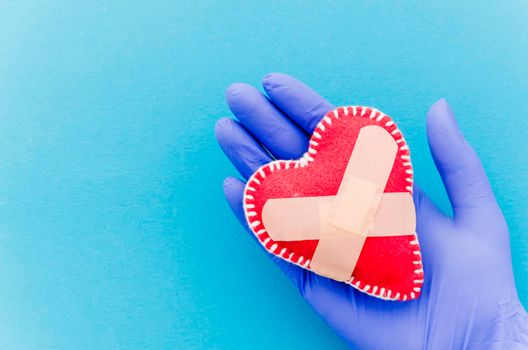 close up hand wearing surgical gloves holding heart shaped stitched textile heart with crossed bandages blue backdrop