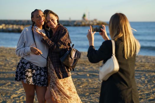 Anonymous woman taking photo of positive multiracial female friends hugging while standing on sandy coast near rippling sea on sunny day