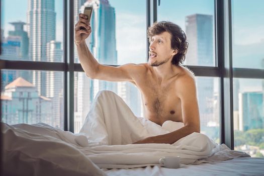 Man wakes up in the morning in an apartment in the downtown area with a view of the skyscrapers and uses smartphone. Life in the noise of the big city concept. Not enough sleep.